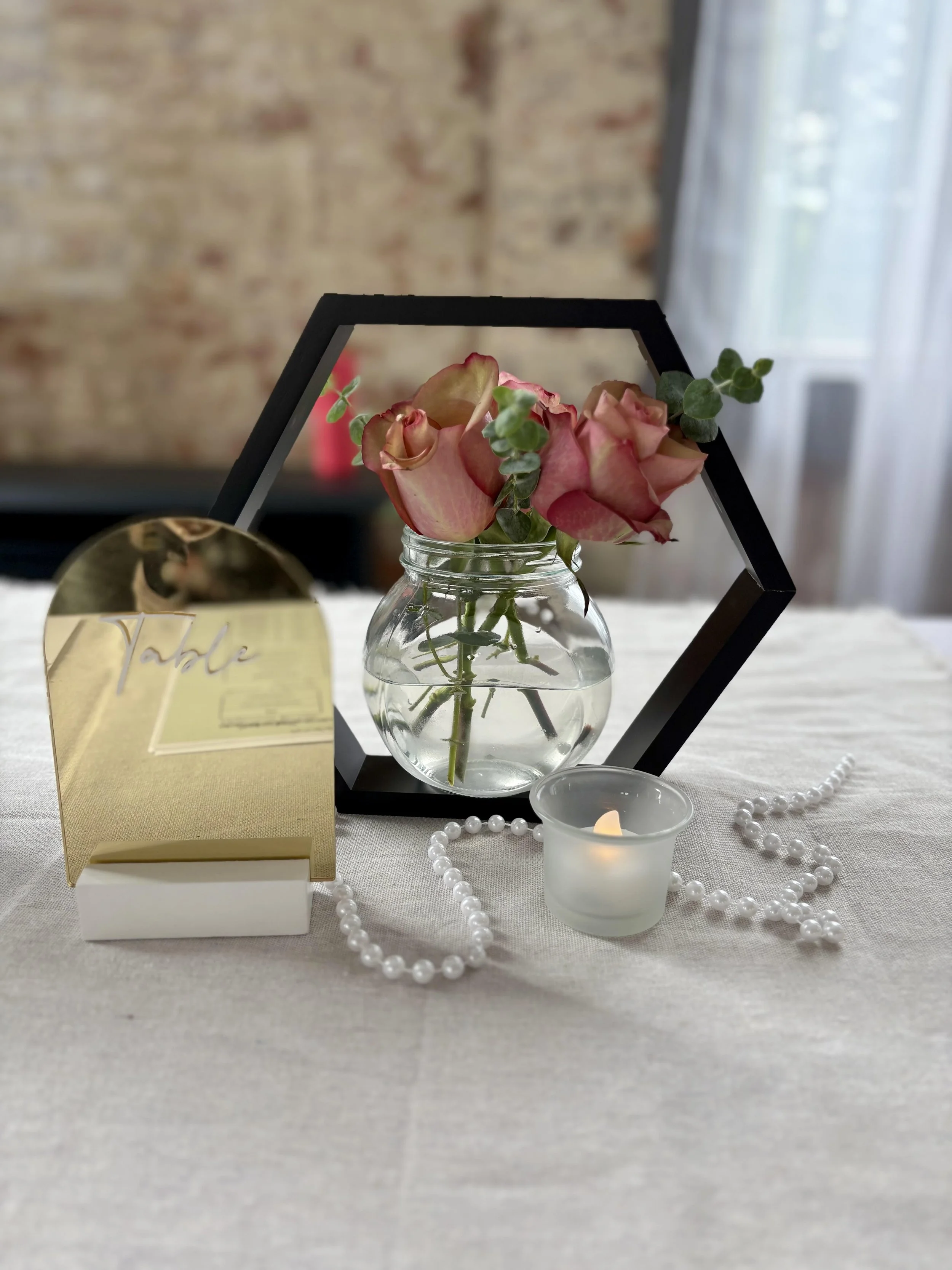 Decorative table centerpiece with pink roses in a glass jar, black geometric frame, small candle, pearl necklace, and a gold mirror with the word 'Table' engraved on it.