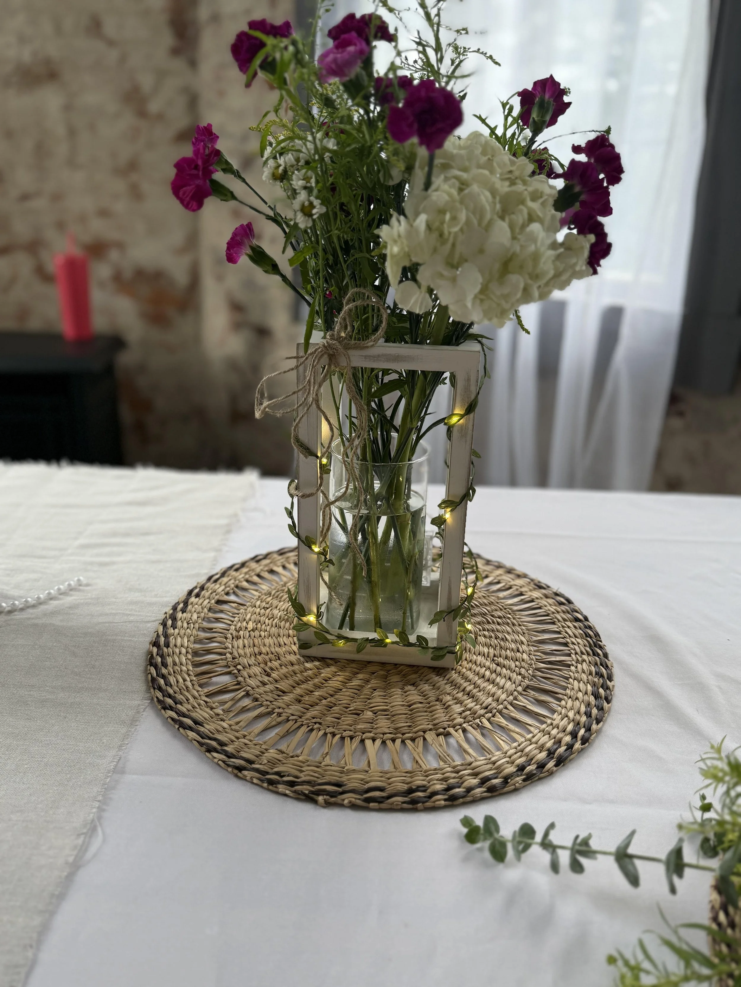 A flower arrangement in a glass vase with purple and white flowers, decorated with string lights, placed on a wicker placemat on a white tablecloth in a room with natural light. Elegant centerpieces at Canton, Ohio Venue. 