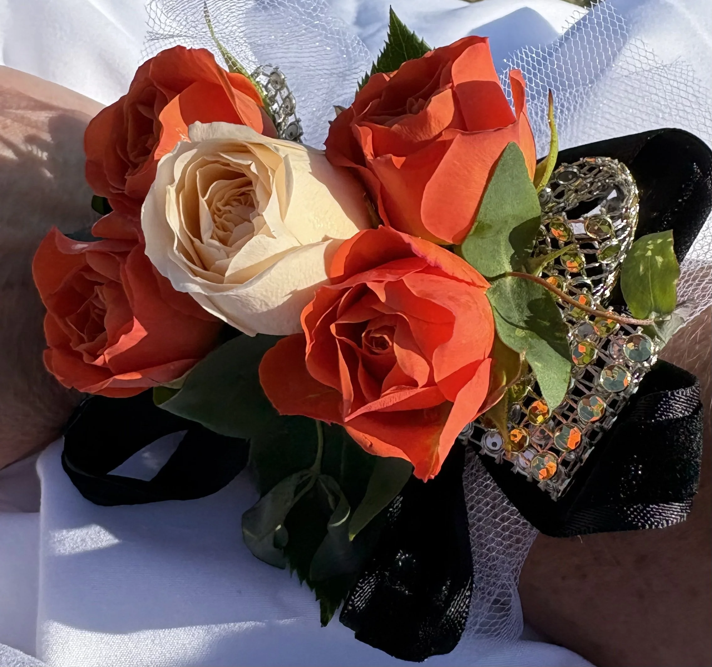 Bouquet of three orange roses and one cream-colored rose, decorated with a sparkly rhinestone band, greenery, black and white ribbons, wrapped in tulle and mesh fabric.