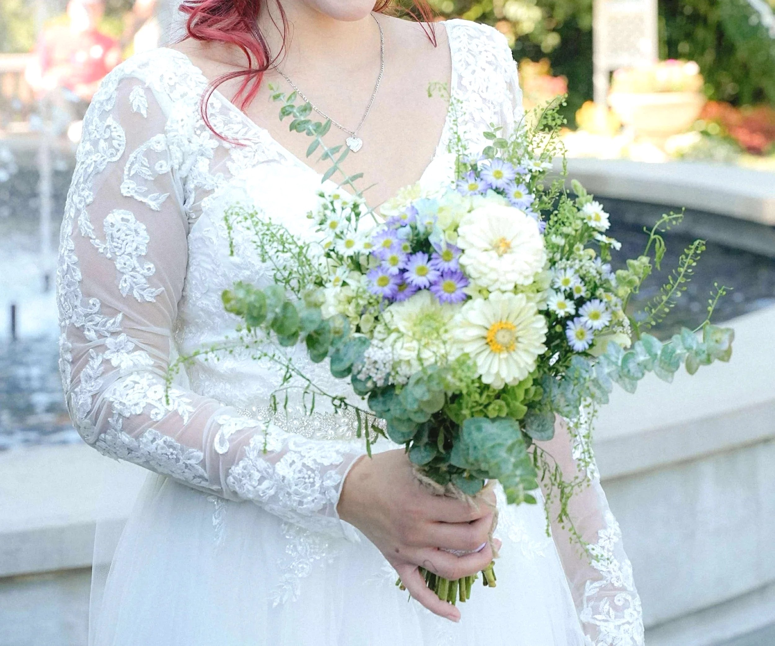 A bride holding a bouquet of white, purple, and green flowers outdoors.