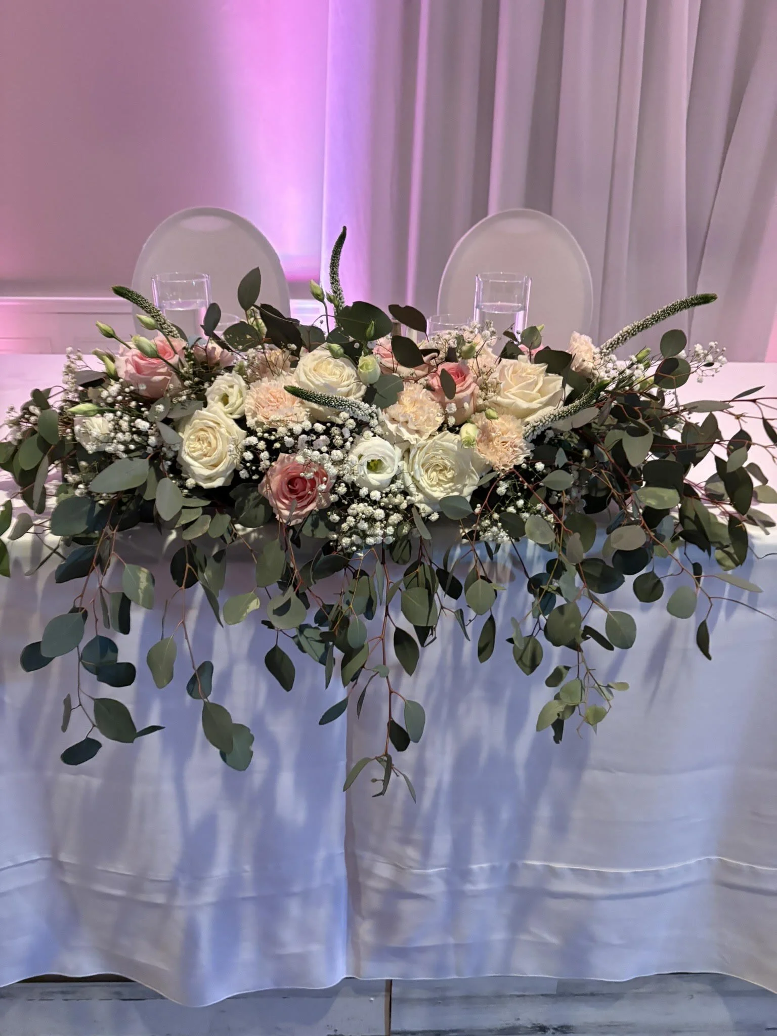 A floral centerpiece on a table with white roses, pink roses, baby's breath, and greenery in front of a white tablecloth, with transparent chairs and pink curtain in the background.