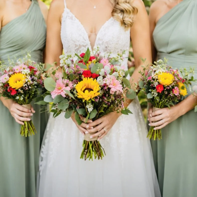 A bride standing between two women, each holding a bouquet. The bride wears a long white lace dress, and the women wear pastel-colored dresses, light green and lavender, all holding small floral arrangements. They are outdoors surrounded by green plants and flowers.