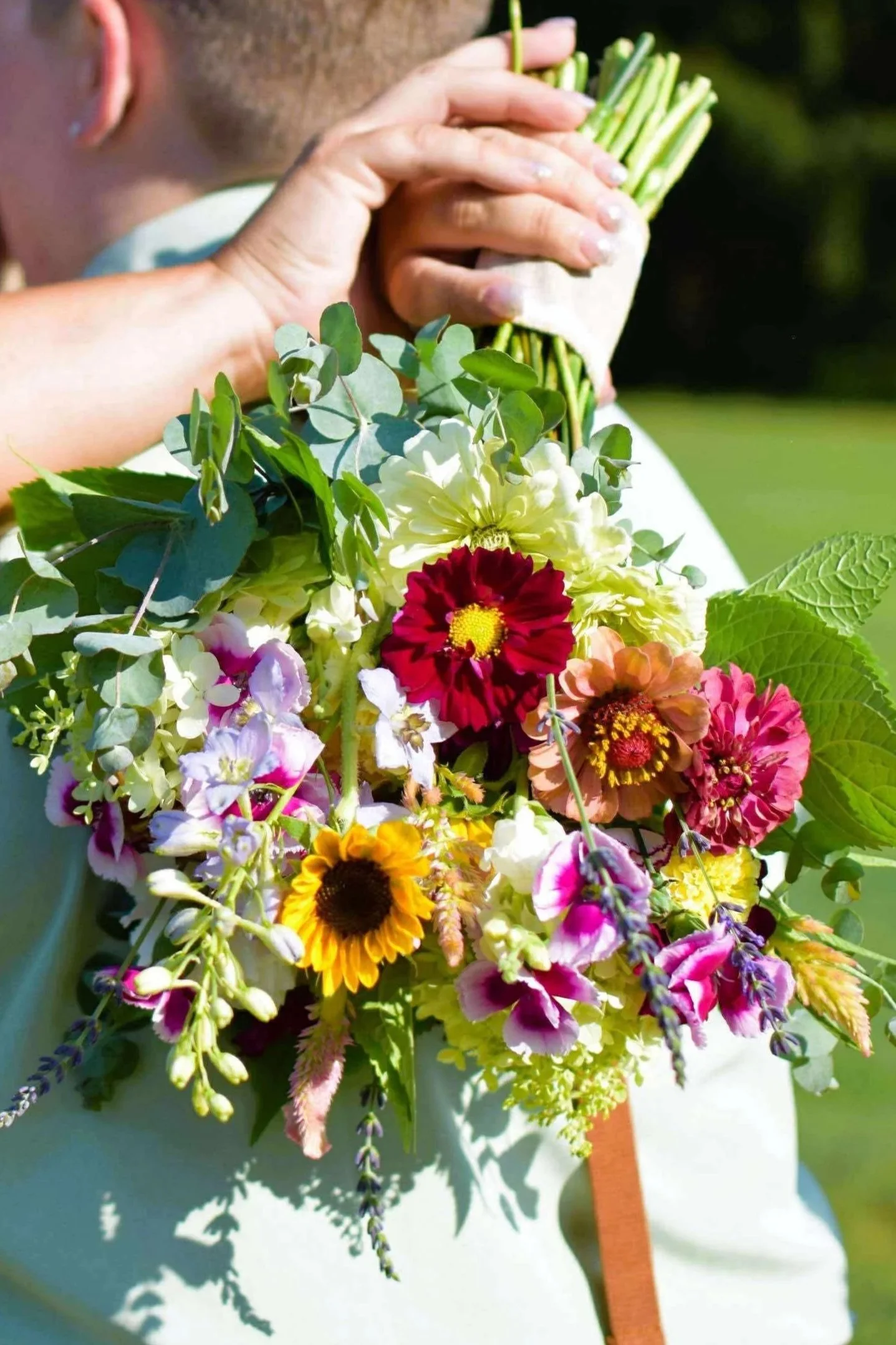 Person holding a bouquet of colorful flowers and green stems outdoors.