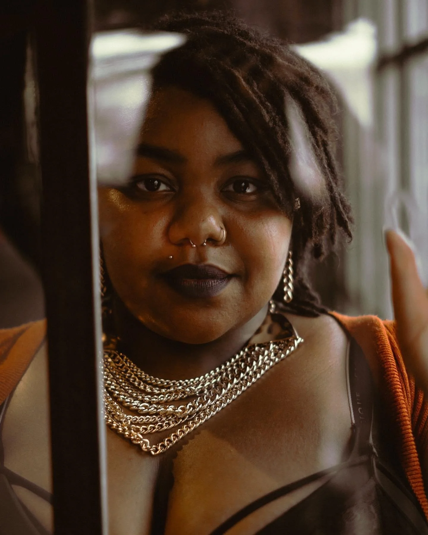 A woman with dark skin and layered jewelry looks through a window, her reflection visible, wearing dark lipstick and a nose ring.