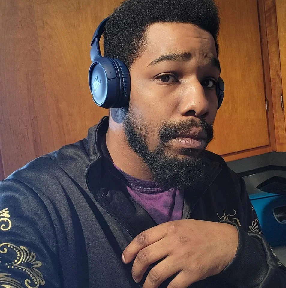 A man with dark curly hair and a beard wearing headphones and a black jacket, looking at the camera with a hand on his chest, in a room with wooden cabinets in the background.
