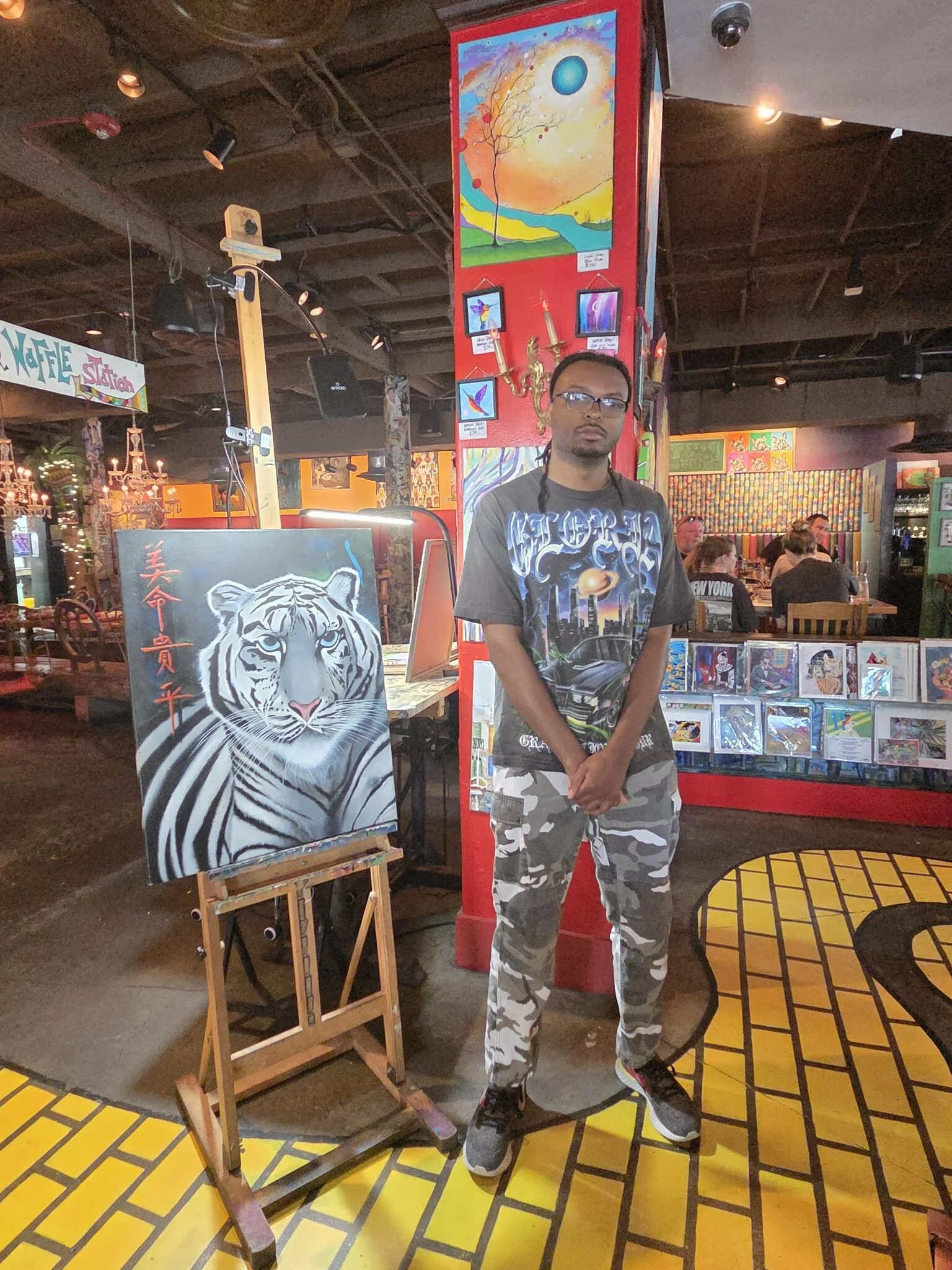 A man standing next to an art display of a tiger painting at an indoor art market or gallery. The background shows colorful artwork and people sitting at tables.