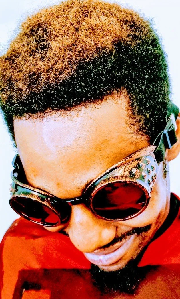 Close-up of a man with two-toned blonde and black hair, wearing dark sunglasses with studs, smiling, and wearing a red shirt.