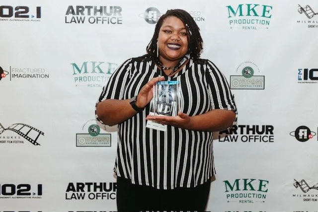 Woman with dreadlocks wearing a black and white striped shirt holding a clear award in front of a backdrop with logos and text.