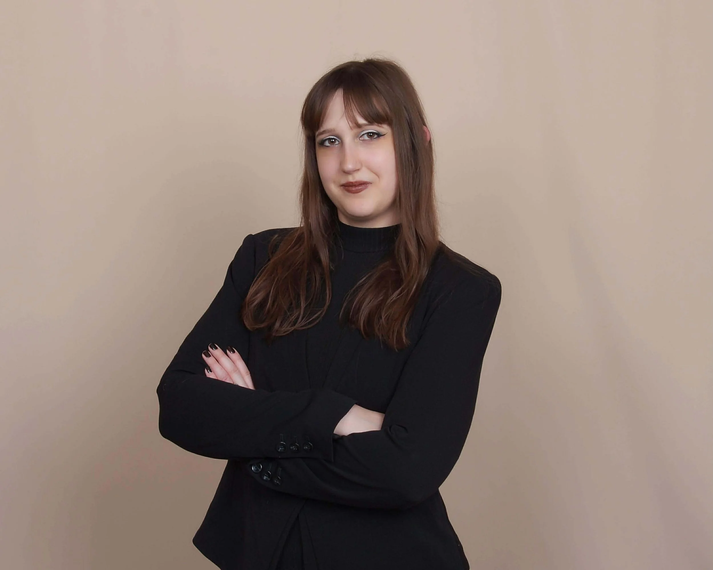 A woman with long brown hair wearing a black blazer and top, standing with arms crossed against a plain beige background.
