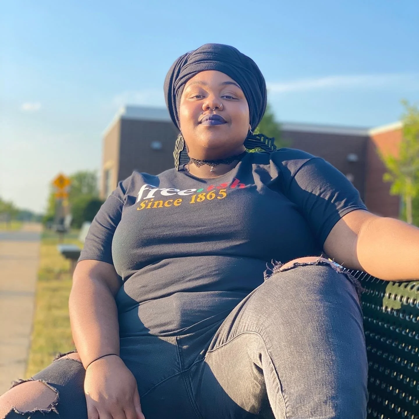 A person sitting on a park bench wearing a black shirt with colorful text and the words 'Since 1865,' black ripped jeans, black headwrap, and jewelry, with a building and trees in the background on a sunny day.