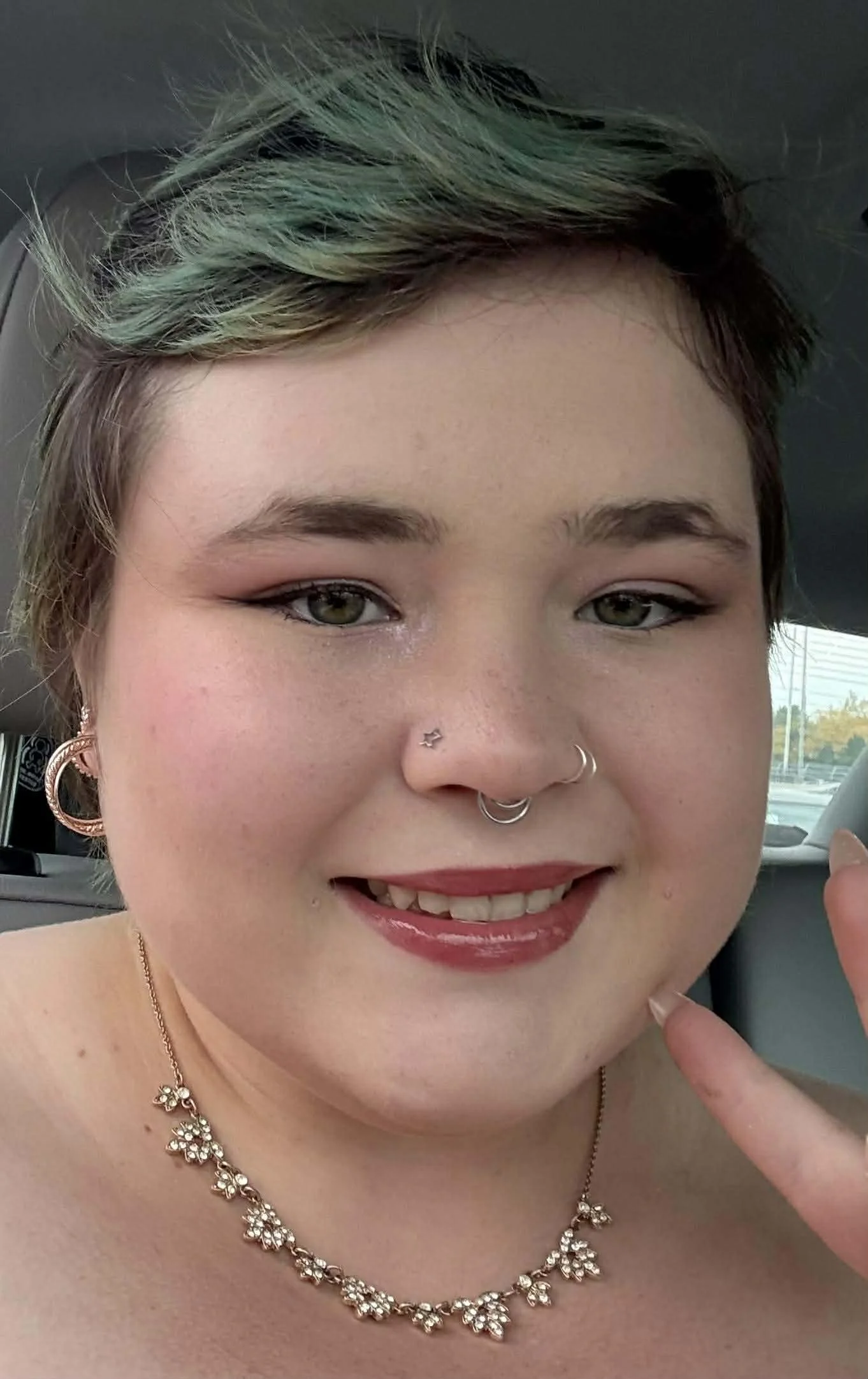 Close-up of a smiling person with short, multicolored hair, multiple facial piercings including a nose ring, a septum ring, and earrings, wearing makeup with lipstick and eyeshadow, a floral necklace, and jewelry on the fingers, inside a car.