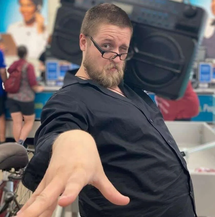A man with glasses and a beard strikes a confident pose with one arm extended in a store, with a large speaker on his shoulder and people shopping in the background.