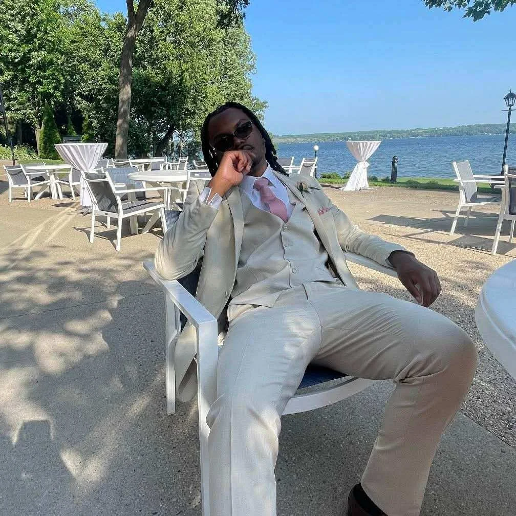 A man dressed in a light-colored suit and pink tie sitting outdoors near a lake, with white chairs and tables around, shade trees, and a clear blue sky.