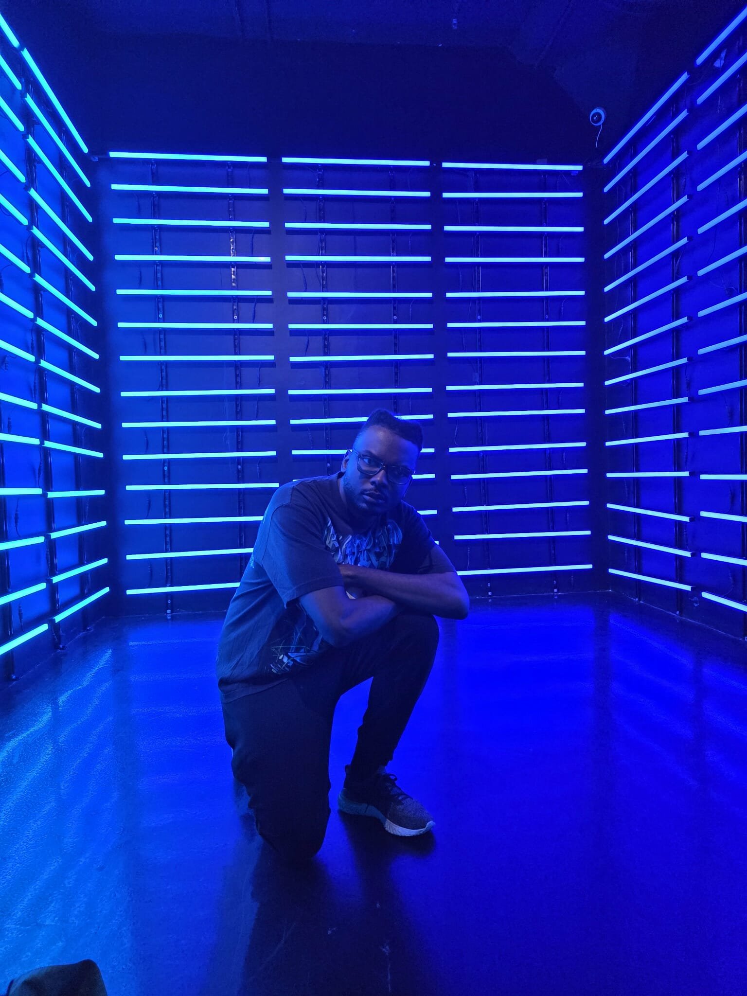 A man kneeling in a room illuminated with blue neon lights on the walls, with his arms crossed and wearing glasses and casual clothes.
