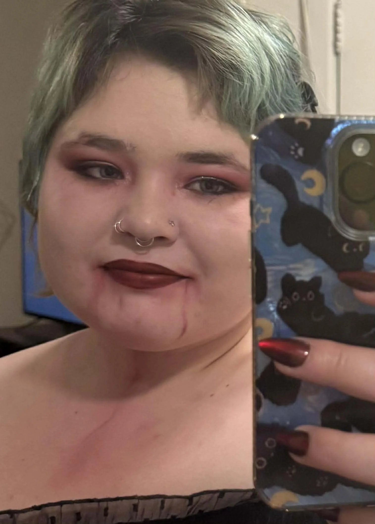 A person with short, multicolored hair, dark lipstick, and makeup, taking a selfie in a mirror with a phone that has a black cat with stars and moons design on the case.