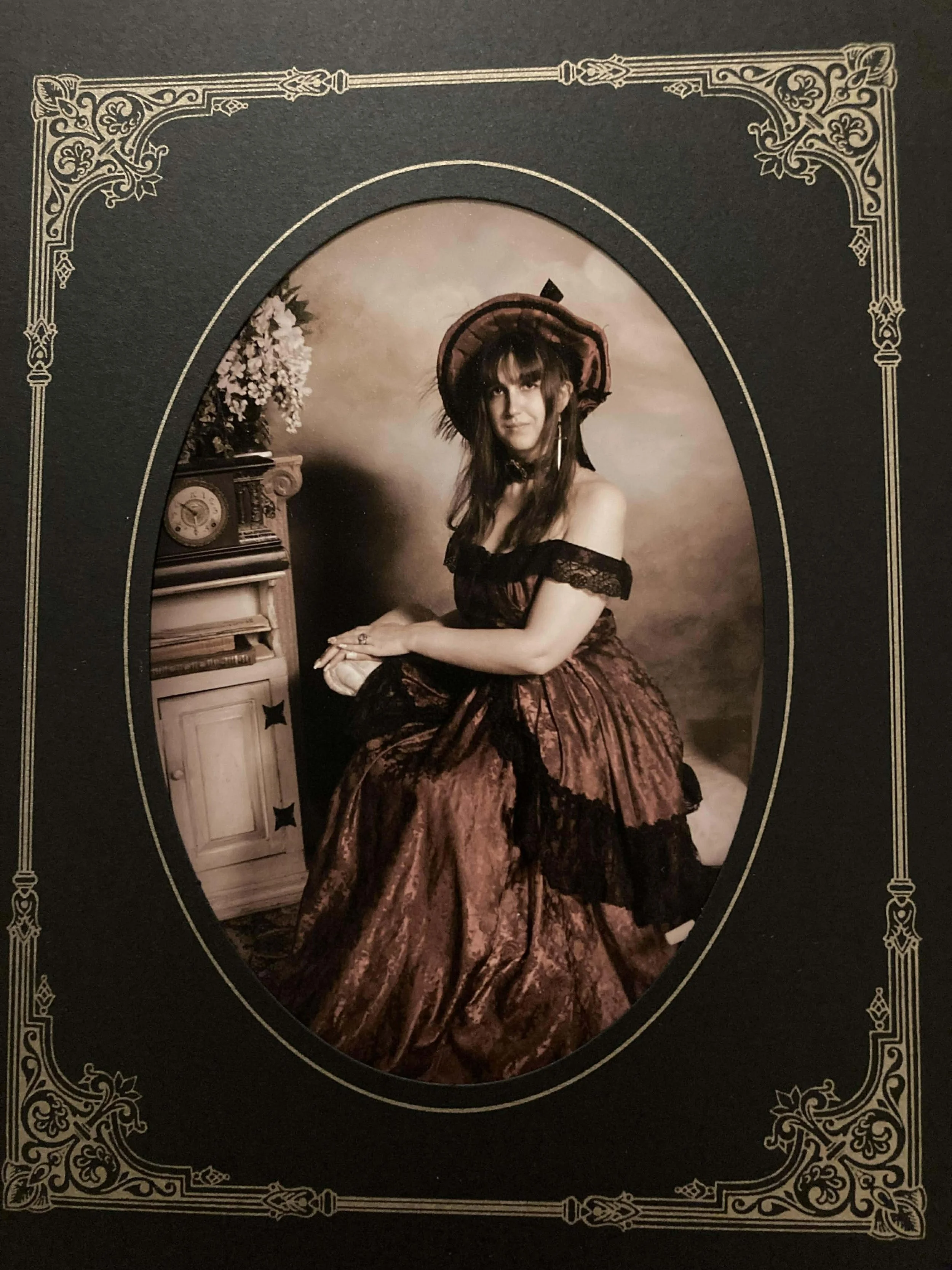 A vintage sepia-toned photograph of a young woman dressed in Victorian-era clothing, sitting beside a small table with a clock and flowers, wearing a wide-brimmed hat and looking at the camera, inside an ornate photo frame.