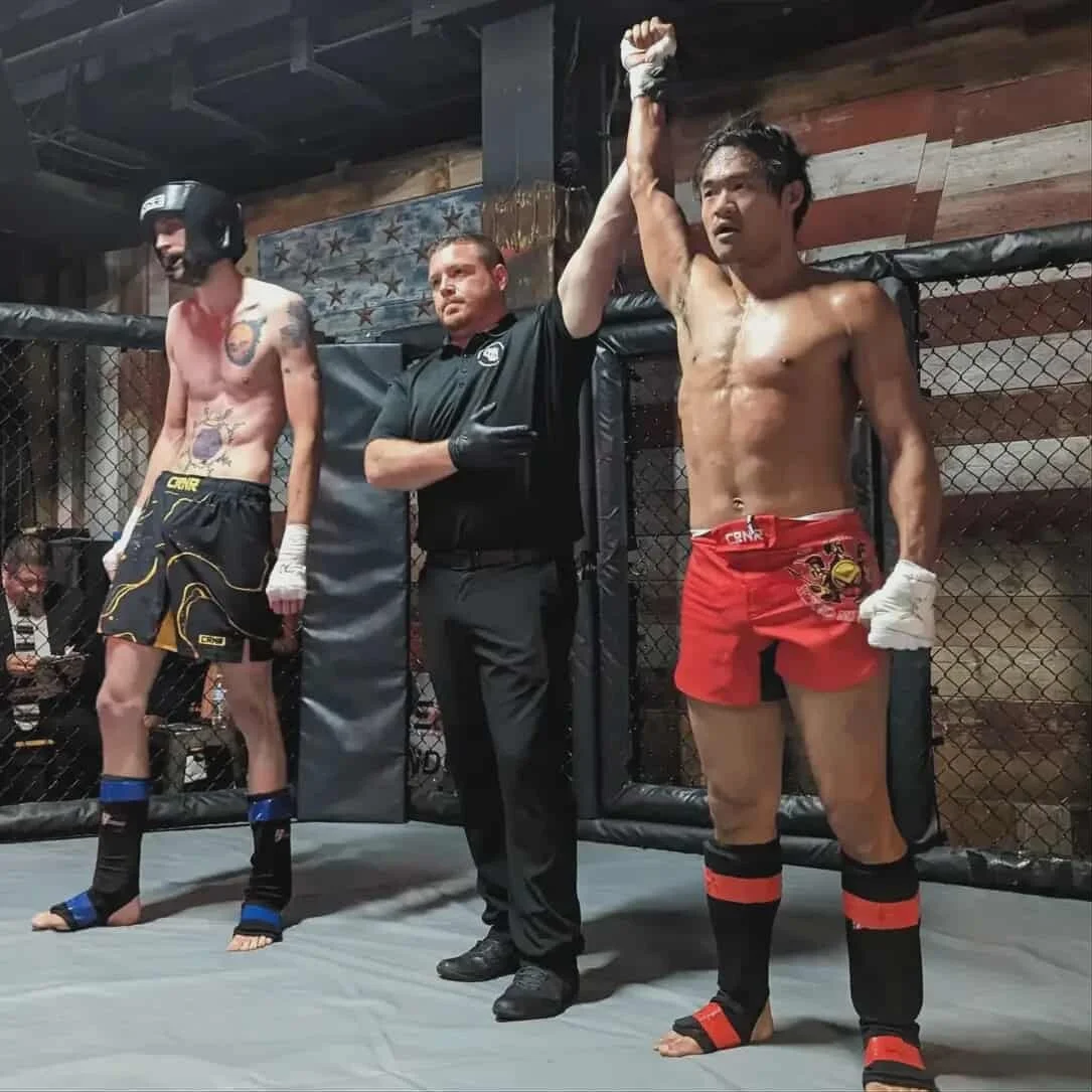 Mixed martial arts fighter with red shorts wins, referee raises his hand, another fighter in black shorts and helmet looks down in the cage.
