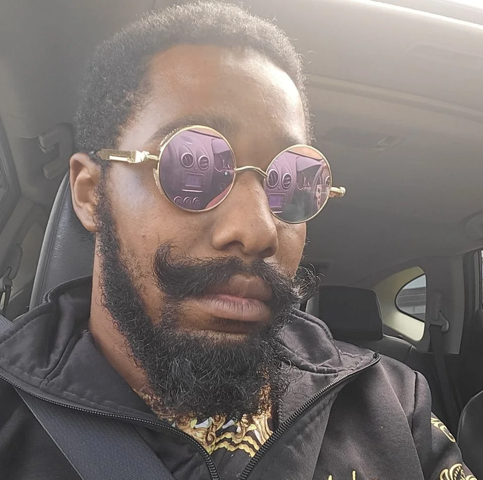 Man with curly hair and a beard wearing round sunglasses with pink reflective lenses, sitting in a car with a seatbelt on.