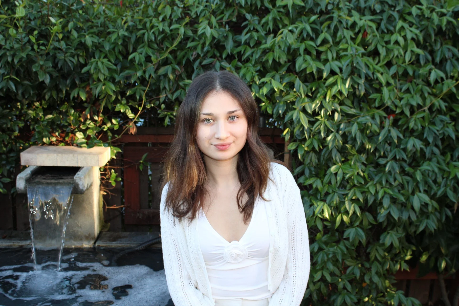 A young woman with shoulder-length brown hair and blue eyes smiling slightly at the camera, standing outdoors in front of green foliage and a small fountain.