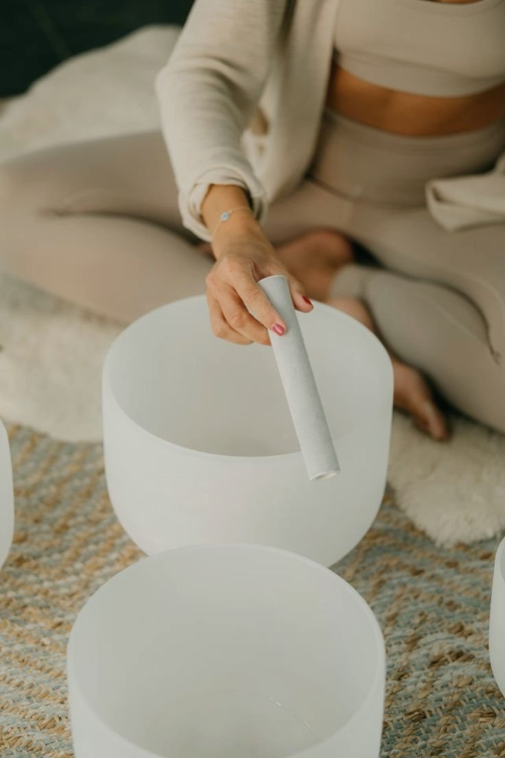 A person sitting cross-legged on a rug, holding a white tube over a white singing bowl. The person is wearing a cream-colored outfit with cutouts and has painted nails.