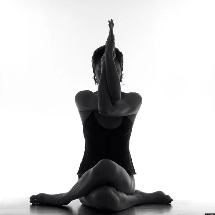A woman with curly hair doing a yoga pose, kneeling on the ground with one leg bent and the other leg extended back. She is smiling and wearing a beige sports bra and matching high-waisted shorts, with her arm raised overhead.