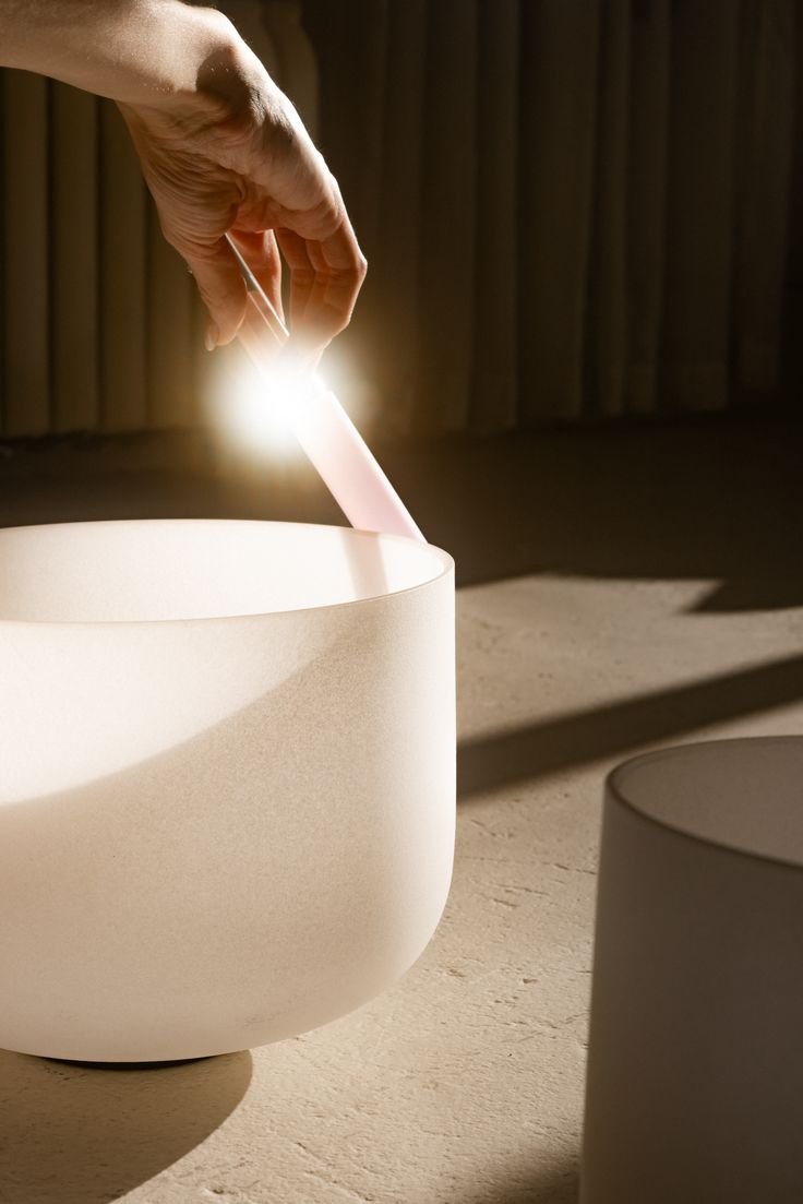 A person sitting cross-legged on a rug, holding a white tube over a white singing bowl. The person is wearing a cream-colored outfit with cutouts and has painted nails.