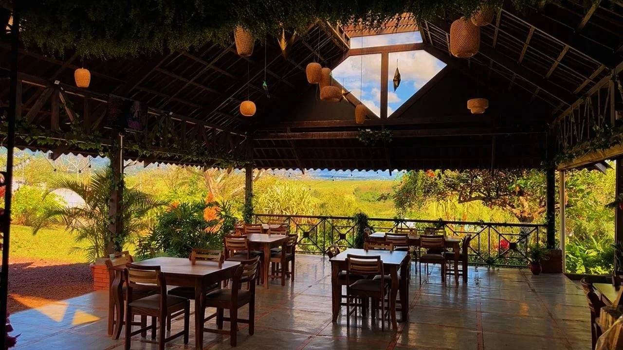 Vista de un comedor al aire libre con mesas y sillas de madera, rodeado de vegetación y un paisaje verde, con un techo de madera y decoraciones en las vigas, y una abertura en el techo que muestra el cielo.