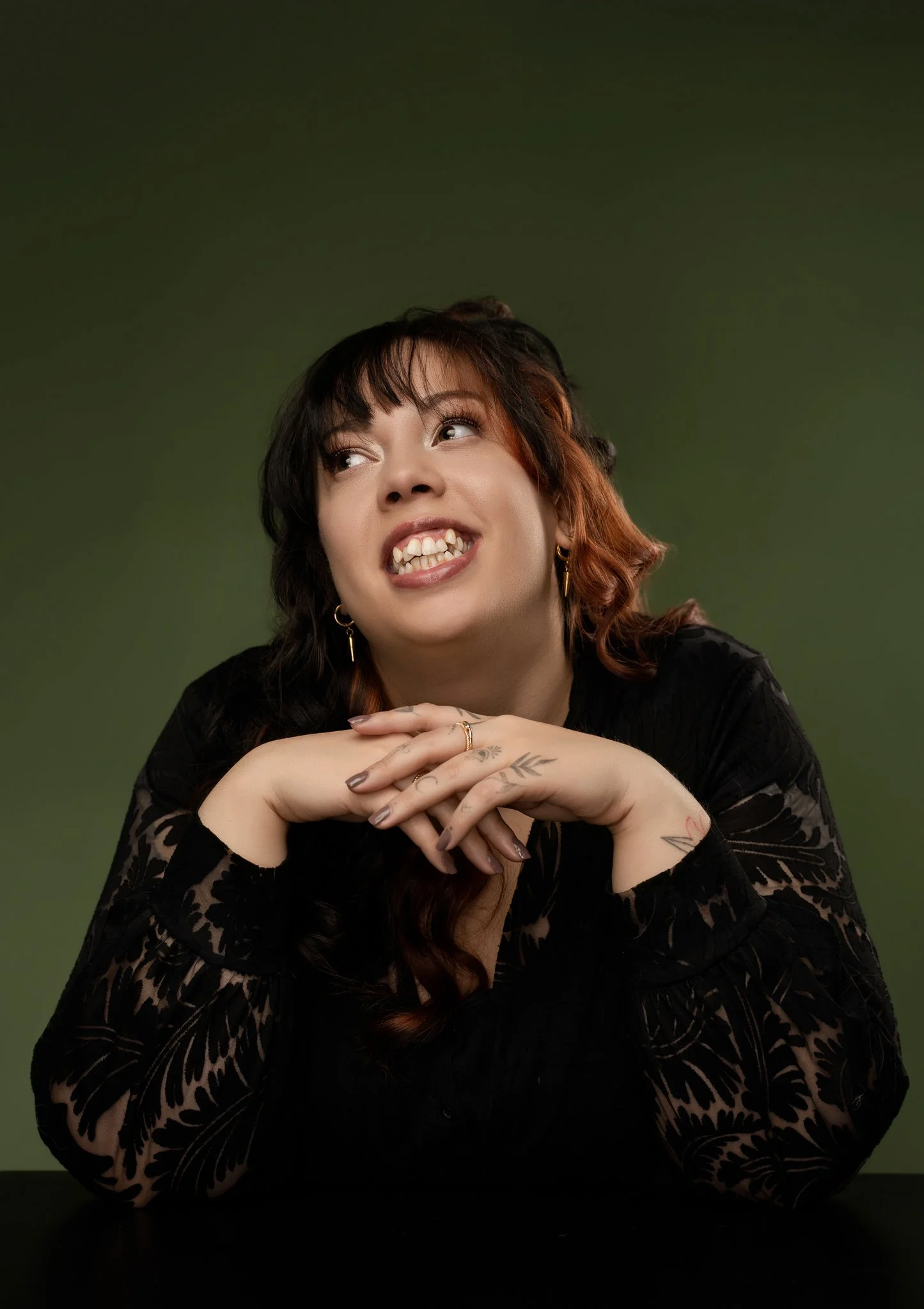 A woman with brown and red hair, wearing a black lace dress and gold earrings, gestures with her hands and has tattoos on her fingers and wrist, against a green background.