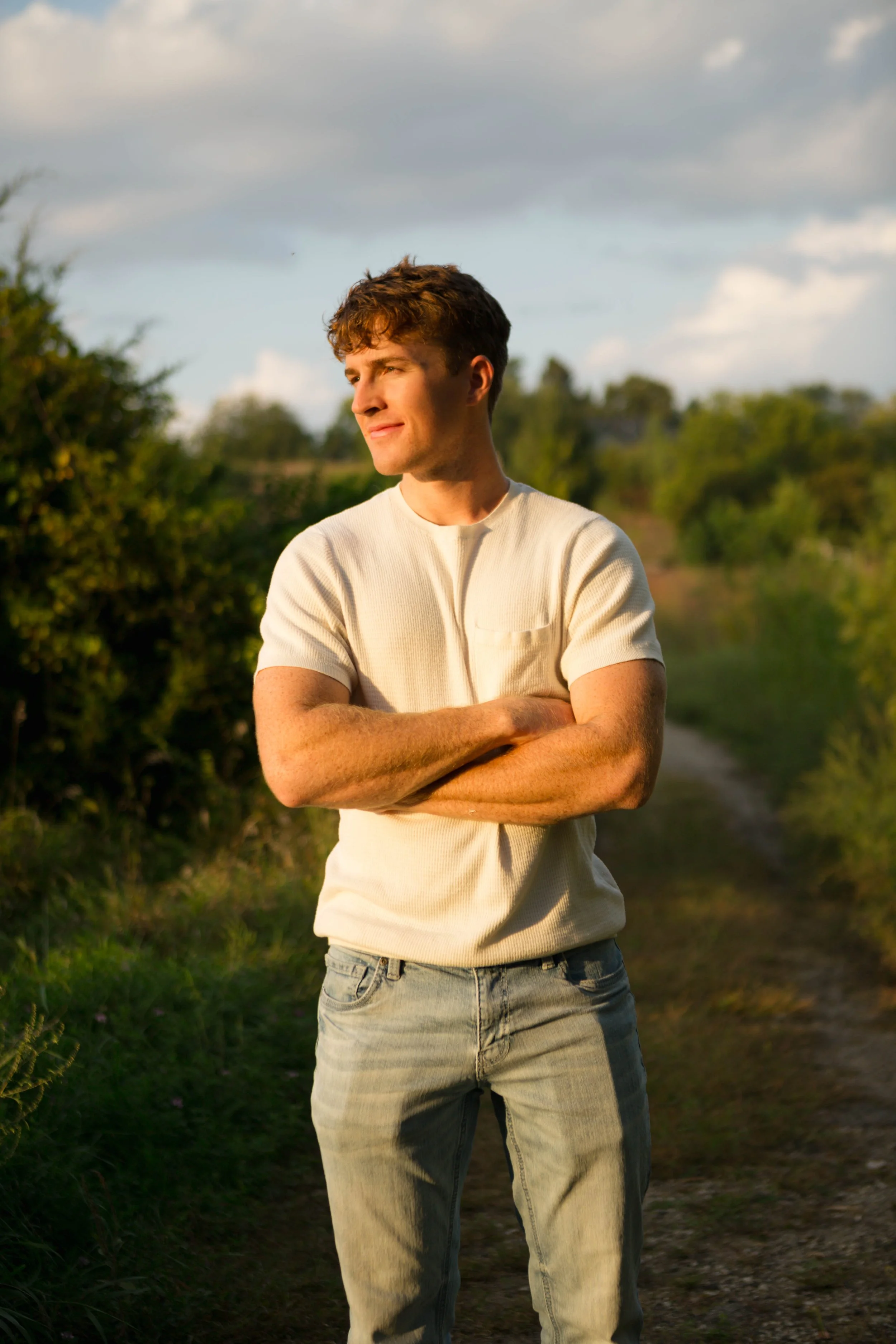 A young man with curly brown hair and light skin standing outdoors on a dirt path, arms crossed, looking to the side, with greenery around and a cloudy sky in the background.