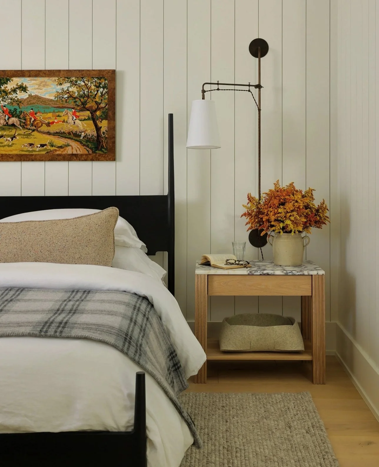 A bedroom with a bed featuring white bedding, a beige pillow, and a gray plaid throw. A nightstand with a marble top holds a vase of orange and yellow flowers, an open book, a glass, and some jewelry. A woven basket is underneath the nightstand. The wall behind is white with vertical wooden paneling. A framed painting of a landscape with people, trees, and animals hangs above the bed. A white pendant light and a black wall lamp are mounted on the wall.