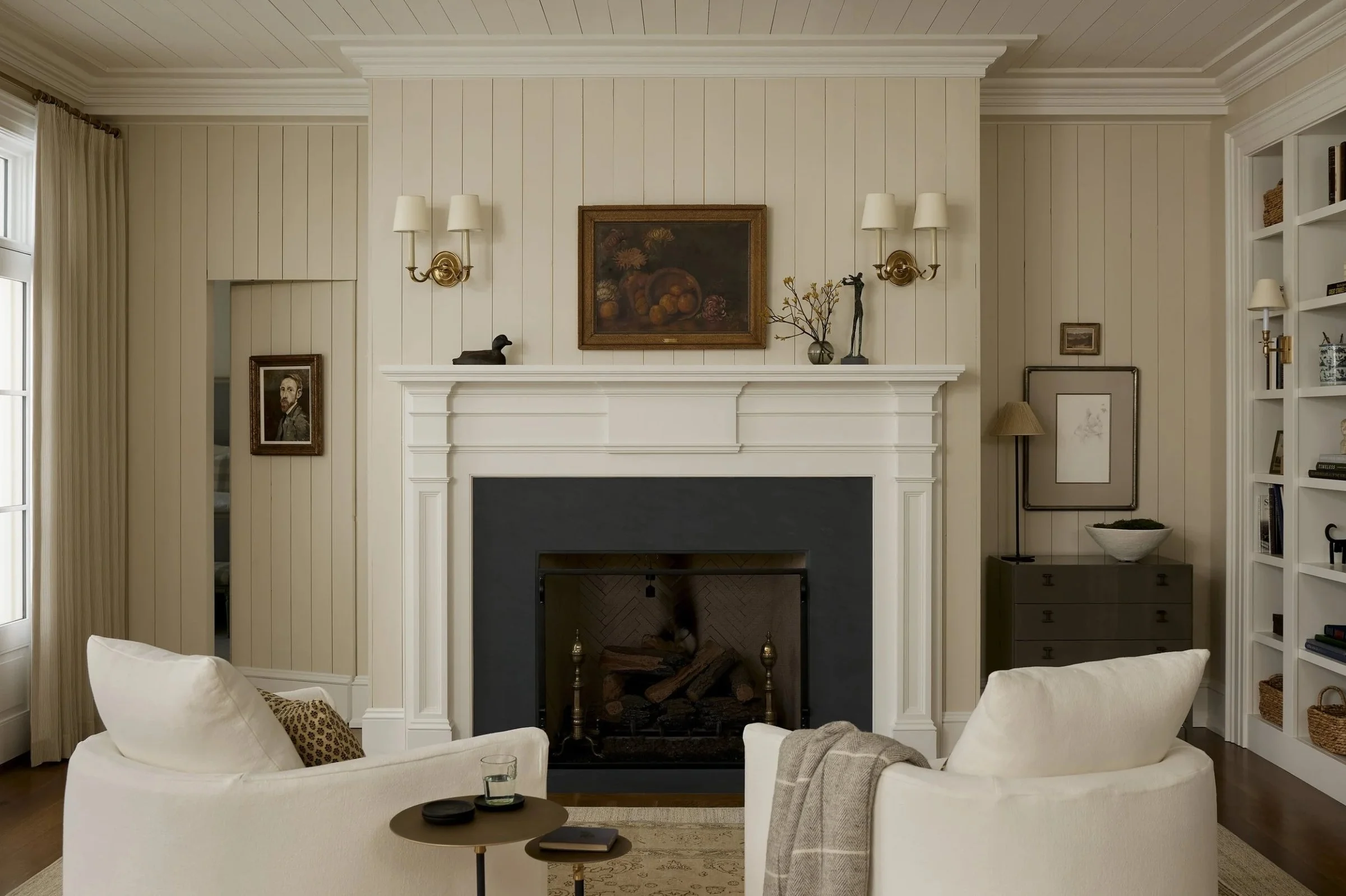 Living room with cream-colored walls and a fireplace. The fireplace has decorative molding and is flanked by wall-mounted lamps. Above it hangs a oil painting of fruit. To the left, there's a window with beige curtains. The room features white sofas with throw pillows, a small round coffee table with a few items, and a built-in bookshelf with decorative baskets and items.