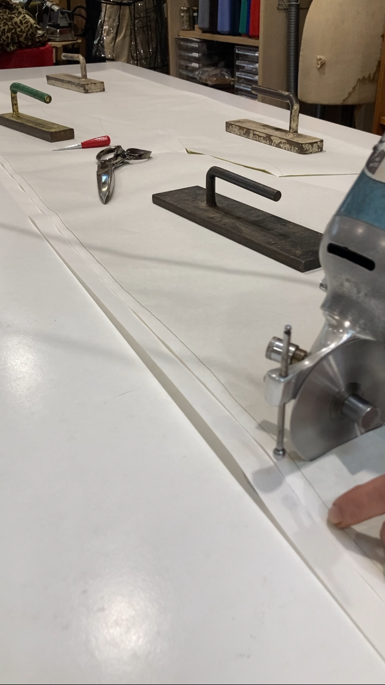 A workbench with cutting tools and equipment, including a rotary cutter, clamps, scissors, and a seam ripper, in a sewing or tailoring workspace.