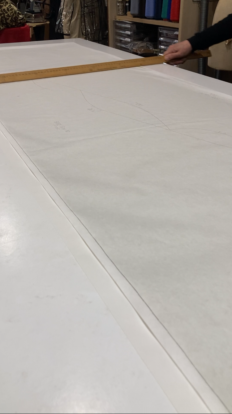 Person using a wooden ruler to measure a large drafting pattern on a white surface in a sewing or tailoring workshop with shelves of fabric and tools in the background.