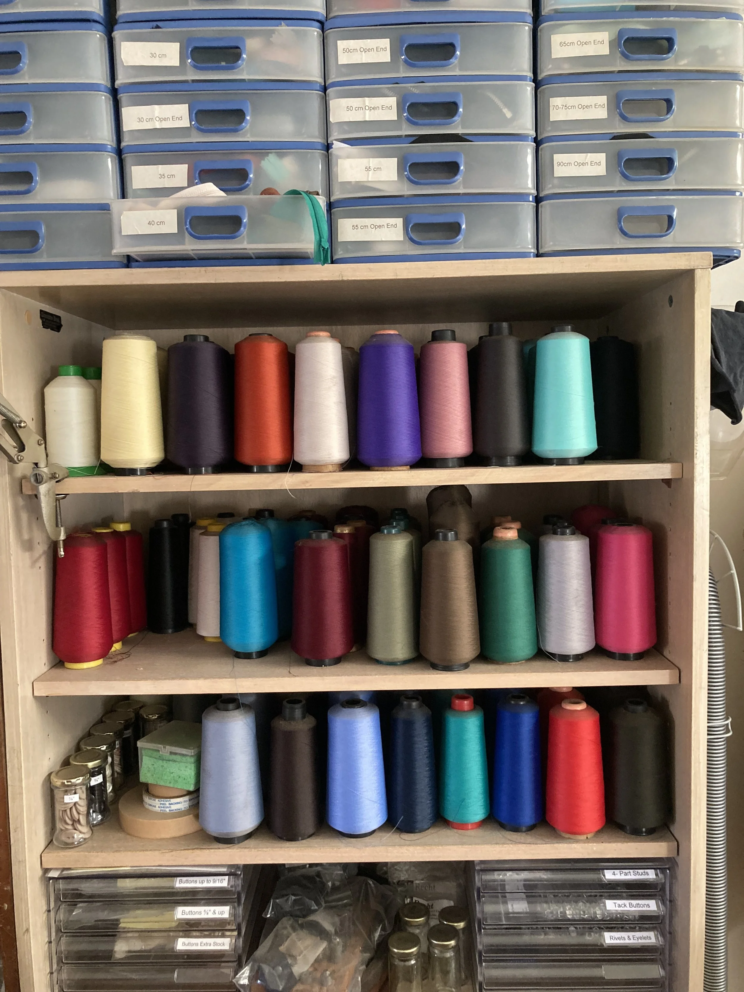 A wooden storage shelf filled with colorful spools of thread and sewing supplies, with plastic containers on top labeled with measurements.
