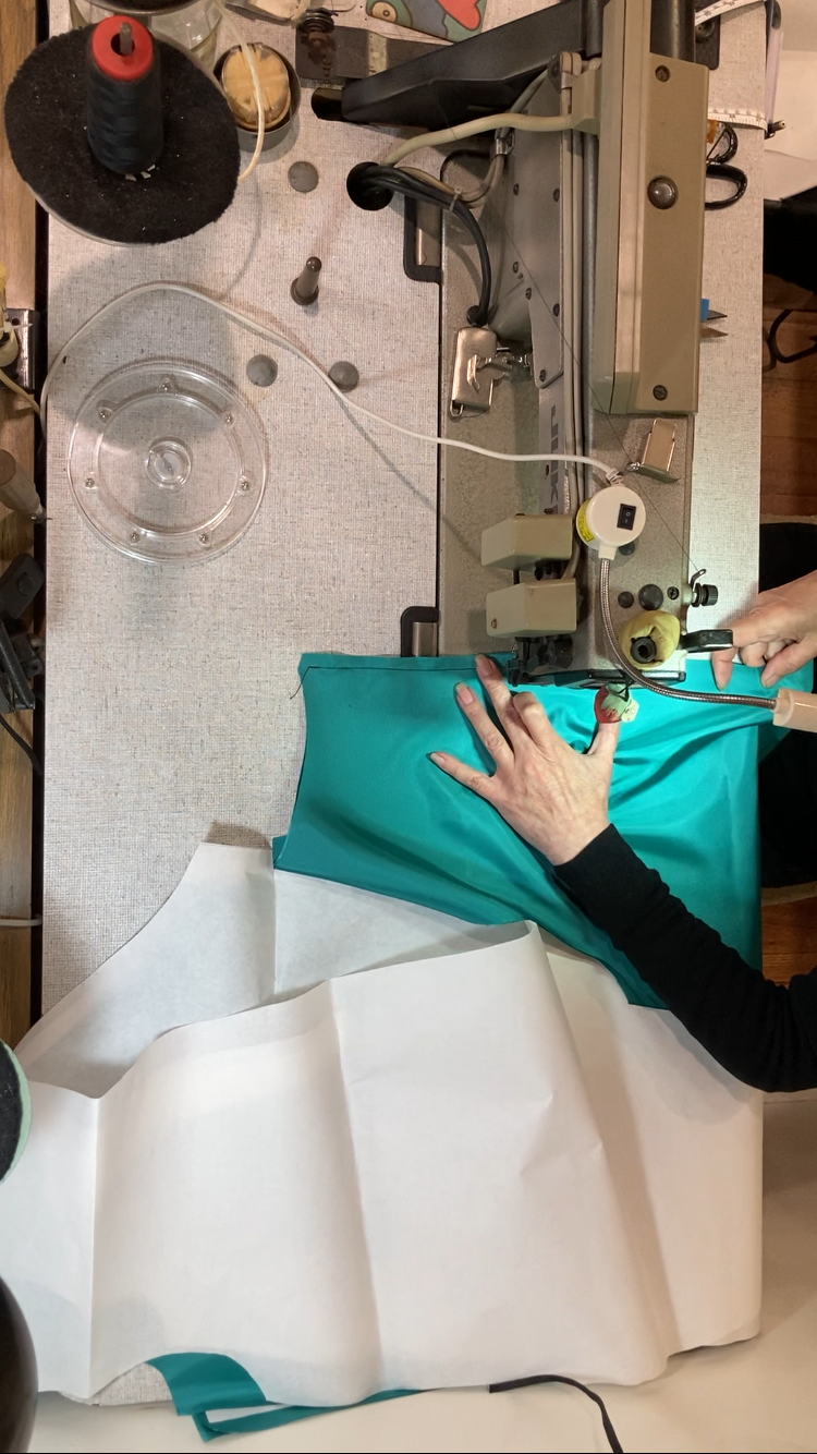 A person sewing turquoise fabric on a vintage industrial sewing machine with white fabric below, surrounded by sewing tools and supplies.