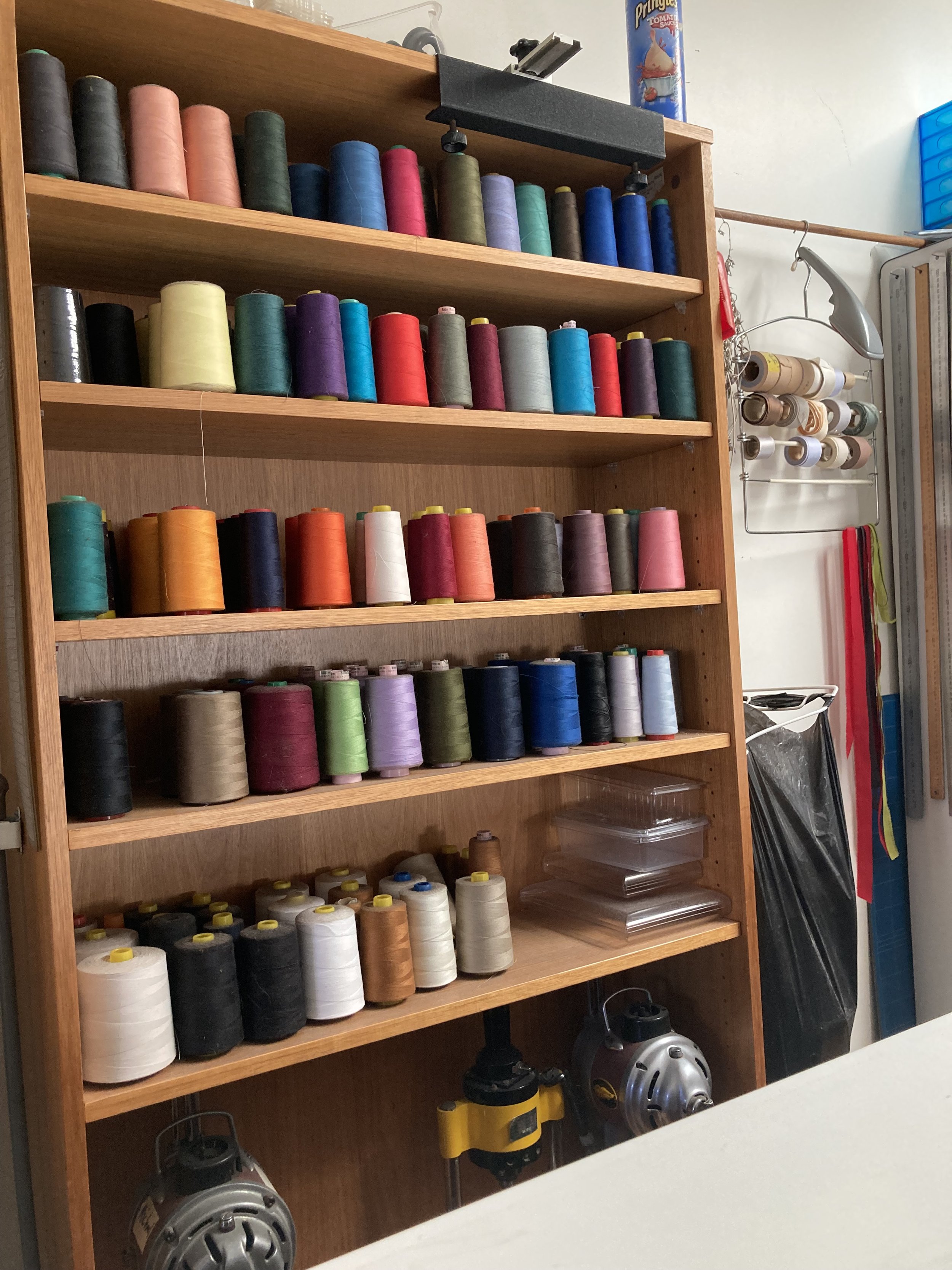 Wooden shelf stocked with colorful spools of thread in various colors, including black, white, red, blue, green, purple, and orange, on multiple shelves in a sewing or craft room.