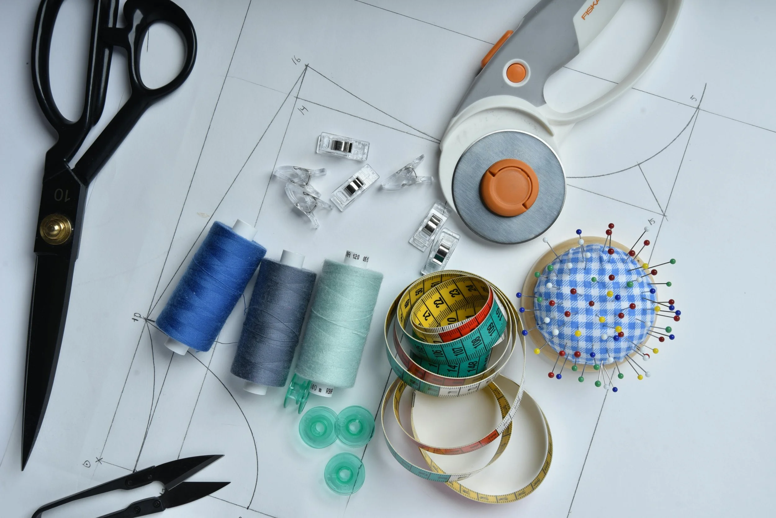 Sewing and tailoring supplies on a pattern paper, including scissors, sewing machine, thread spools, tape measures, pins, and clips.