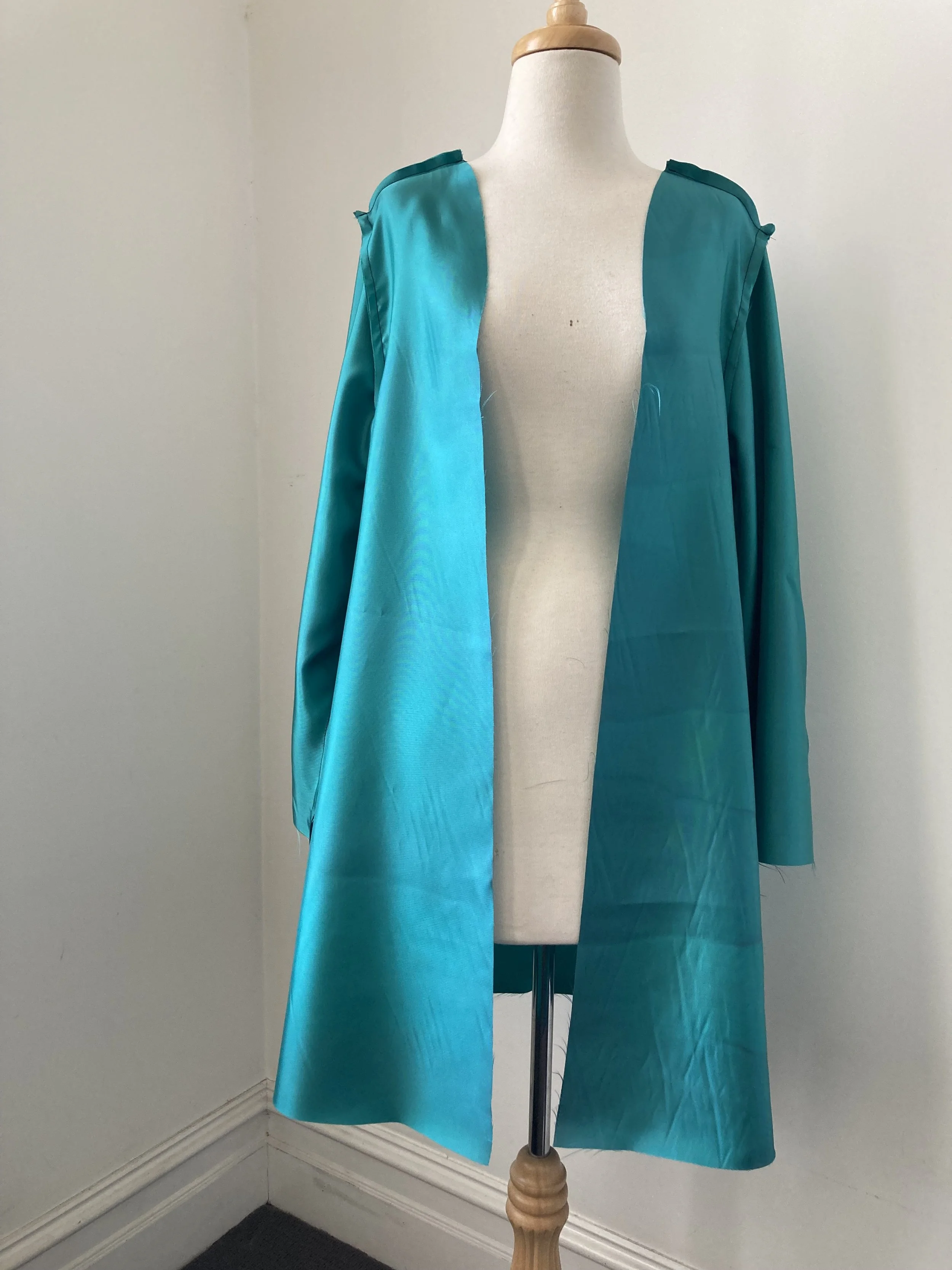 A mannequin draped in a bright turquoise satin fabric, with a long open-front coat or dress, positioned against a plain white wall.