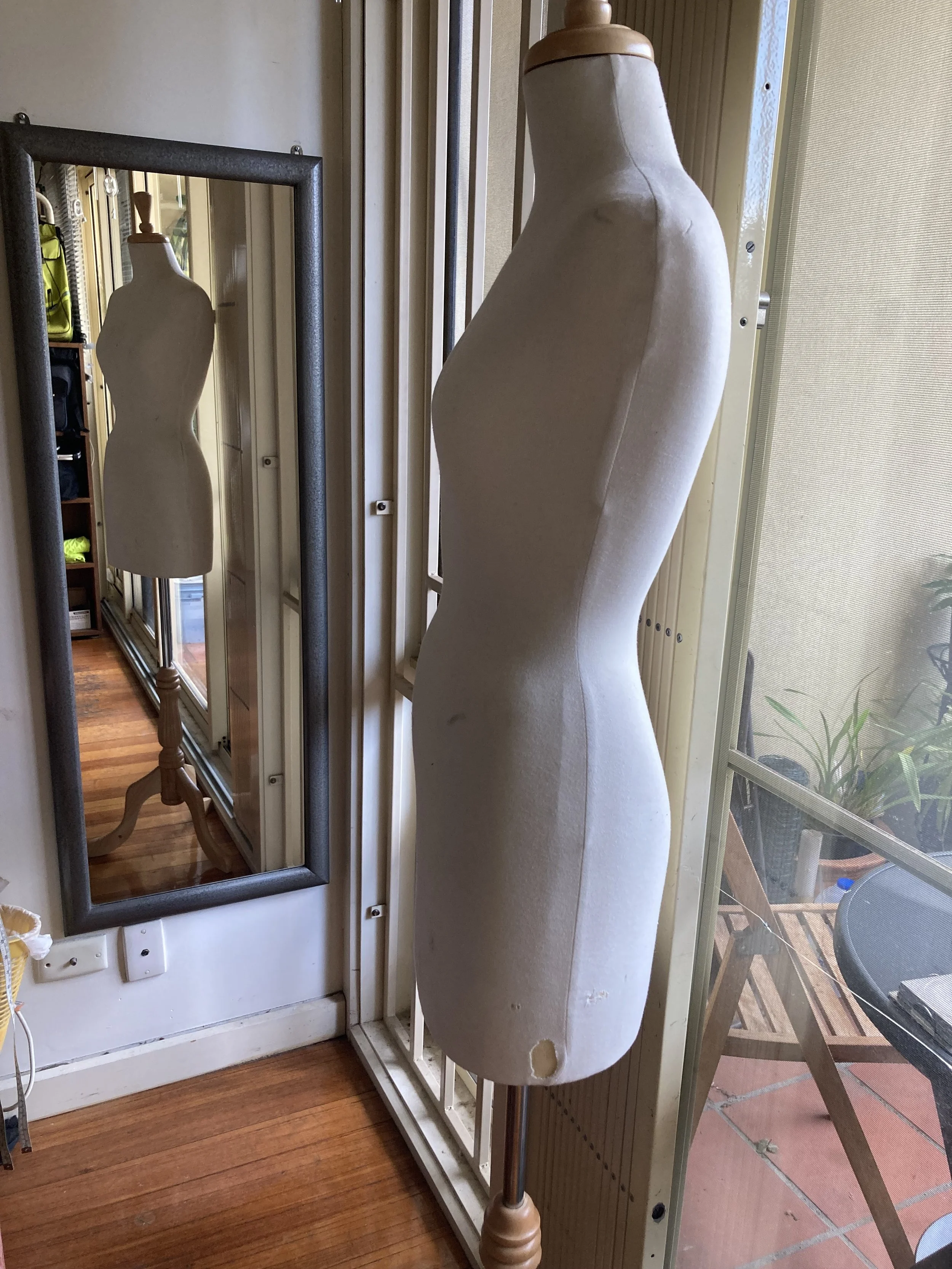 A dress form mannequin standing indoors with a mirror reflecting its front view and a sliding glass door leading to an outdoor patio area.