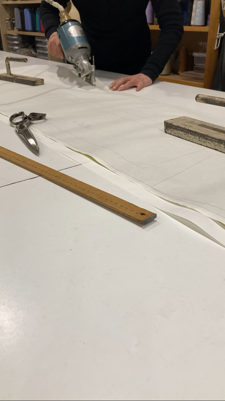 Person cutting fabric with a rotary cutter on a large drafting paper, surrounded by scissors, rulers, and other tools on a workshop table.
