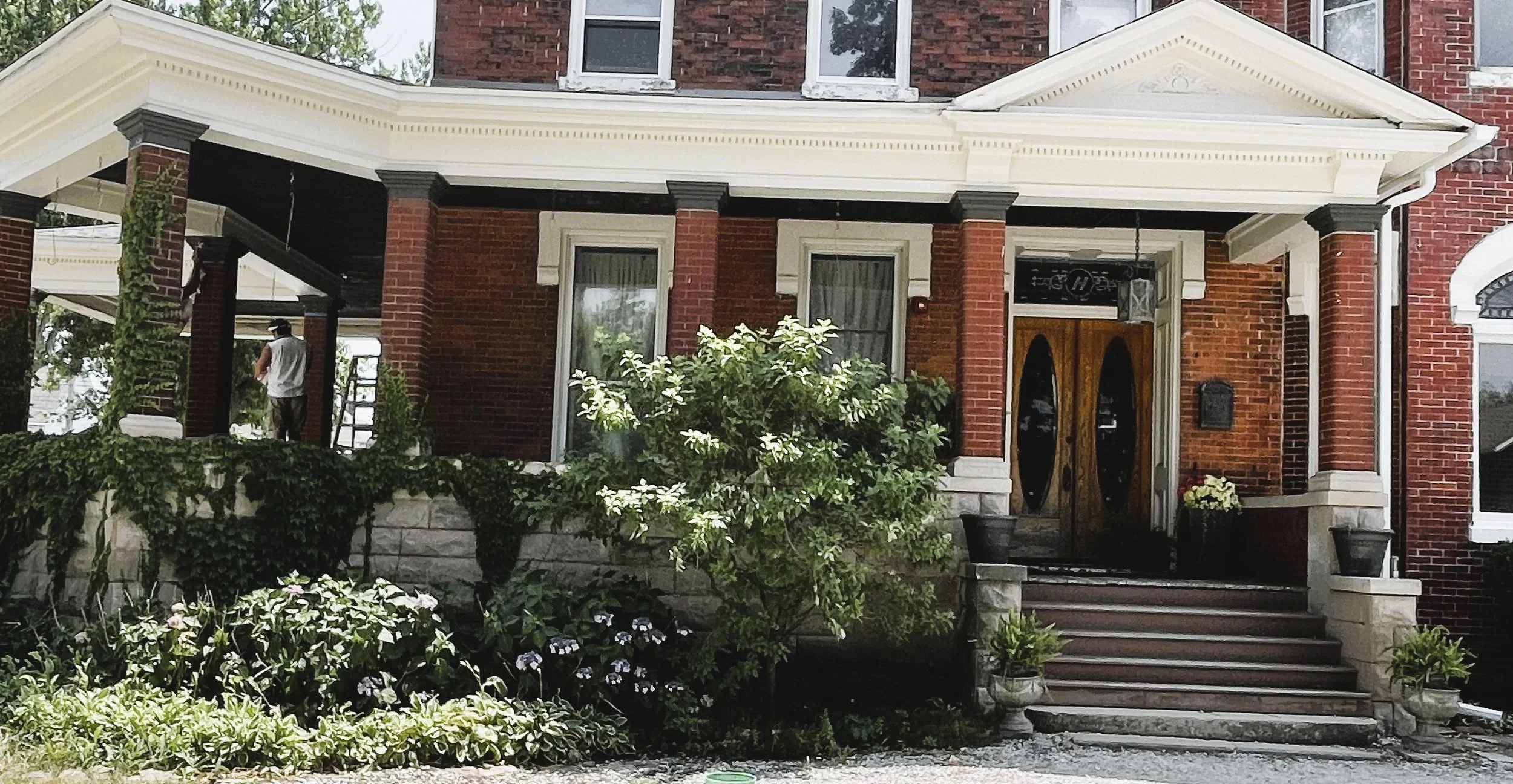 Front view of a two-story brick house with white trim, a porch with steps, and plants and flowers in the garden.