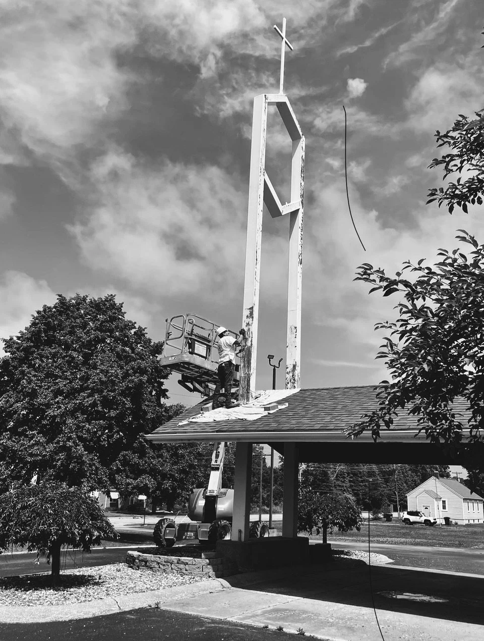 A worker on a lift working on a tall church cross frame near the roof of a building with trees and a house in the background.