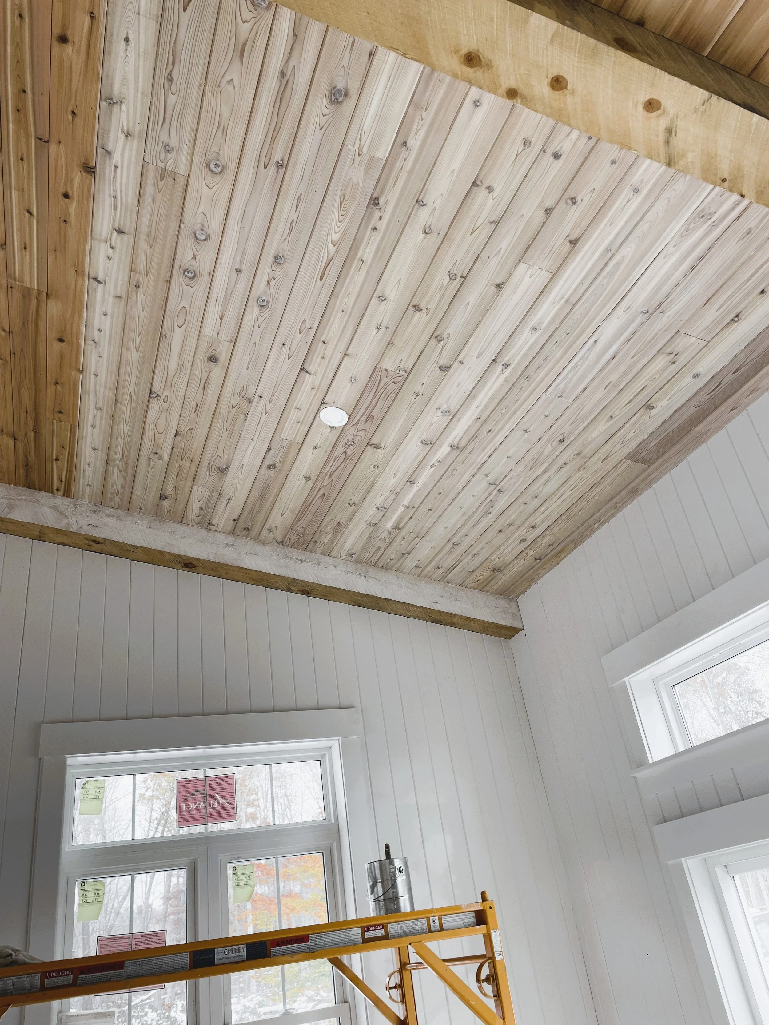 Interior view of a room under construction, showing a wooden ceiling, white paneled walls, a window, and a yellow scaffolding.
