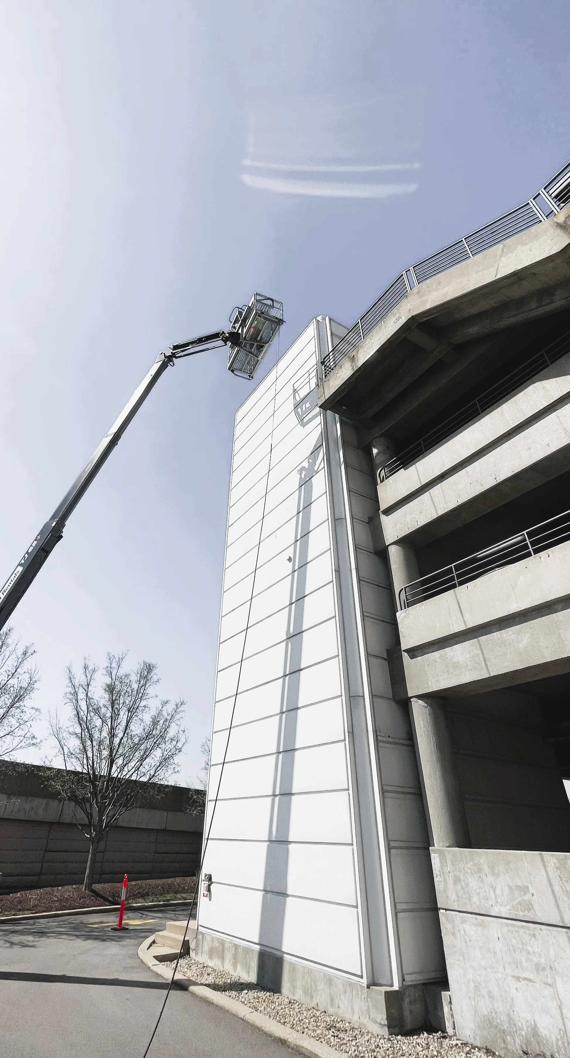 A construction lift with a worker platform extended to the side of a tall building under construction, with a concrete parking garage structure to the right, leafless trees in the background, and an overcast sky.