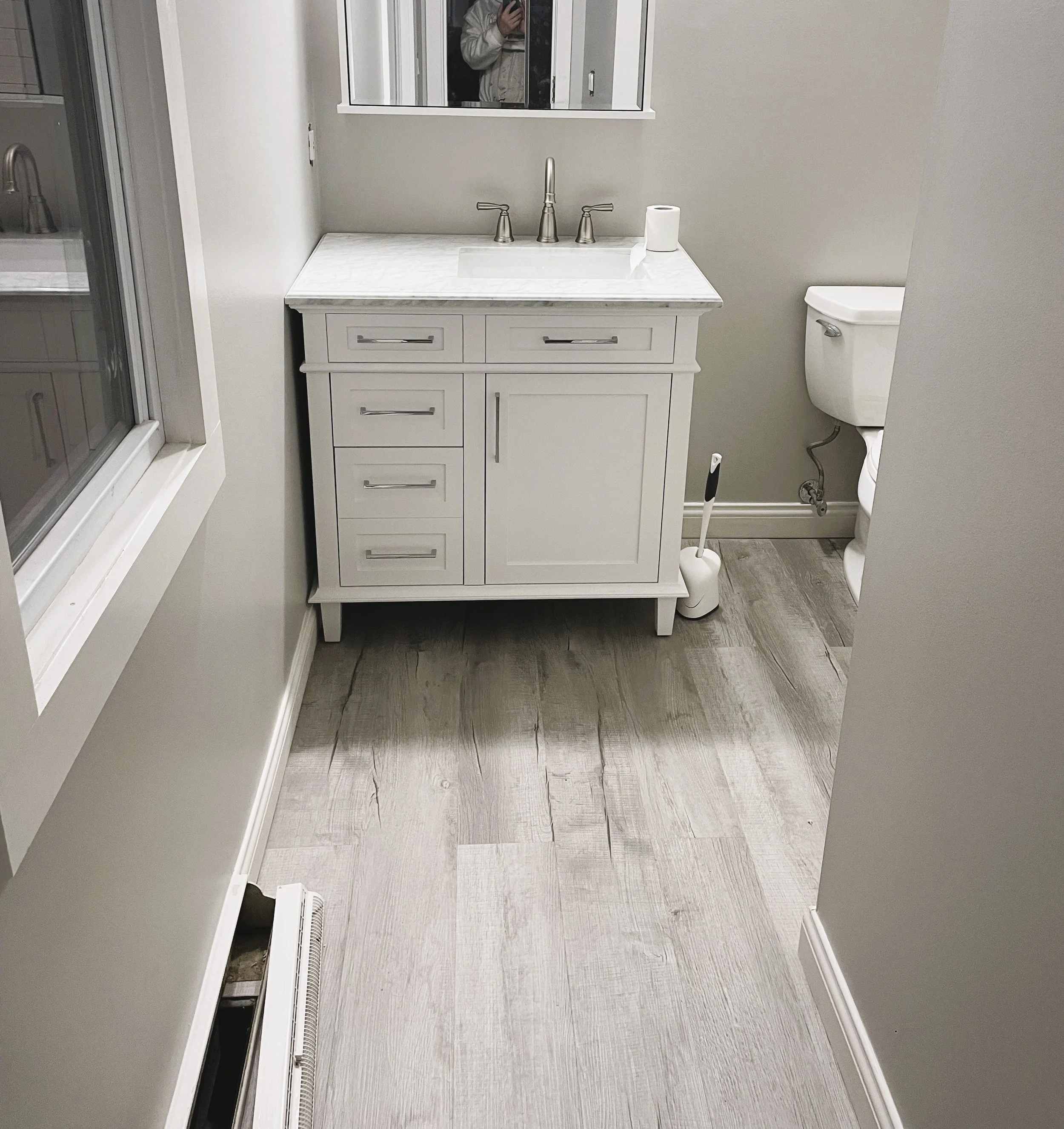 A modern bathroom with a white vanity, marble countertop, and a mirror. There is a toilet to the right and a window on the left. The floor is wood-like tile, and a toilet brush is next to the toilet.