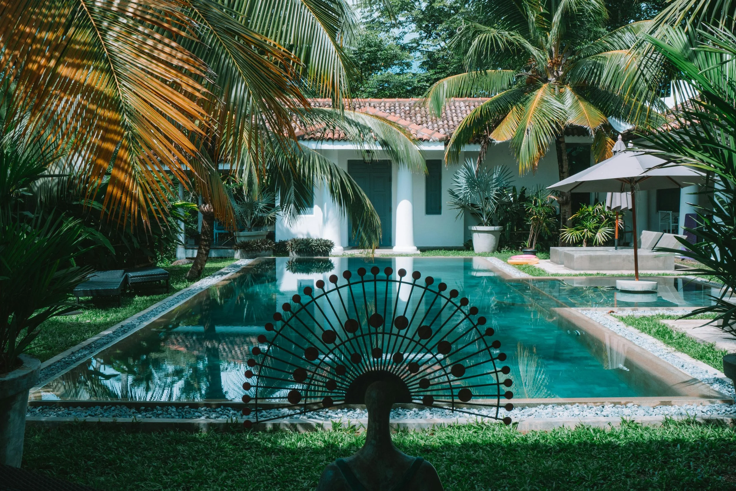 Une villa tropicale avec une piscine, entourée de palmiers et de plantes luxuriantes, sous un ciel bleu.