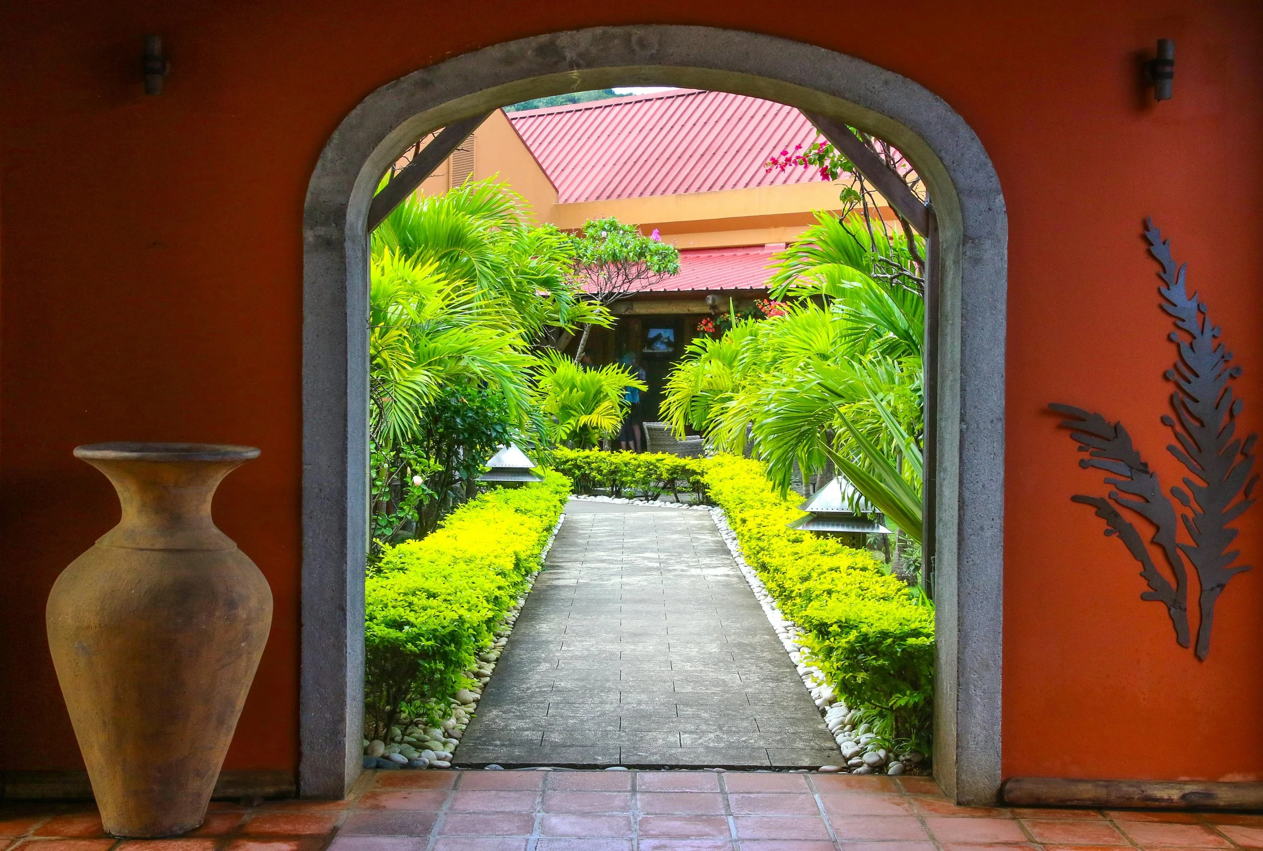 Vue d'un passage dans un jardin tropical vue depuis une ouverture dans un mur orange. Le chemin en pierre mène à une maison avec un toit en tôle rouge, entourée d'arbustes et de palmiers verts. À gauche, un grand vase en terre cuite est placé sur le 