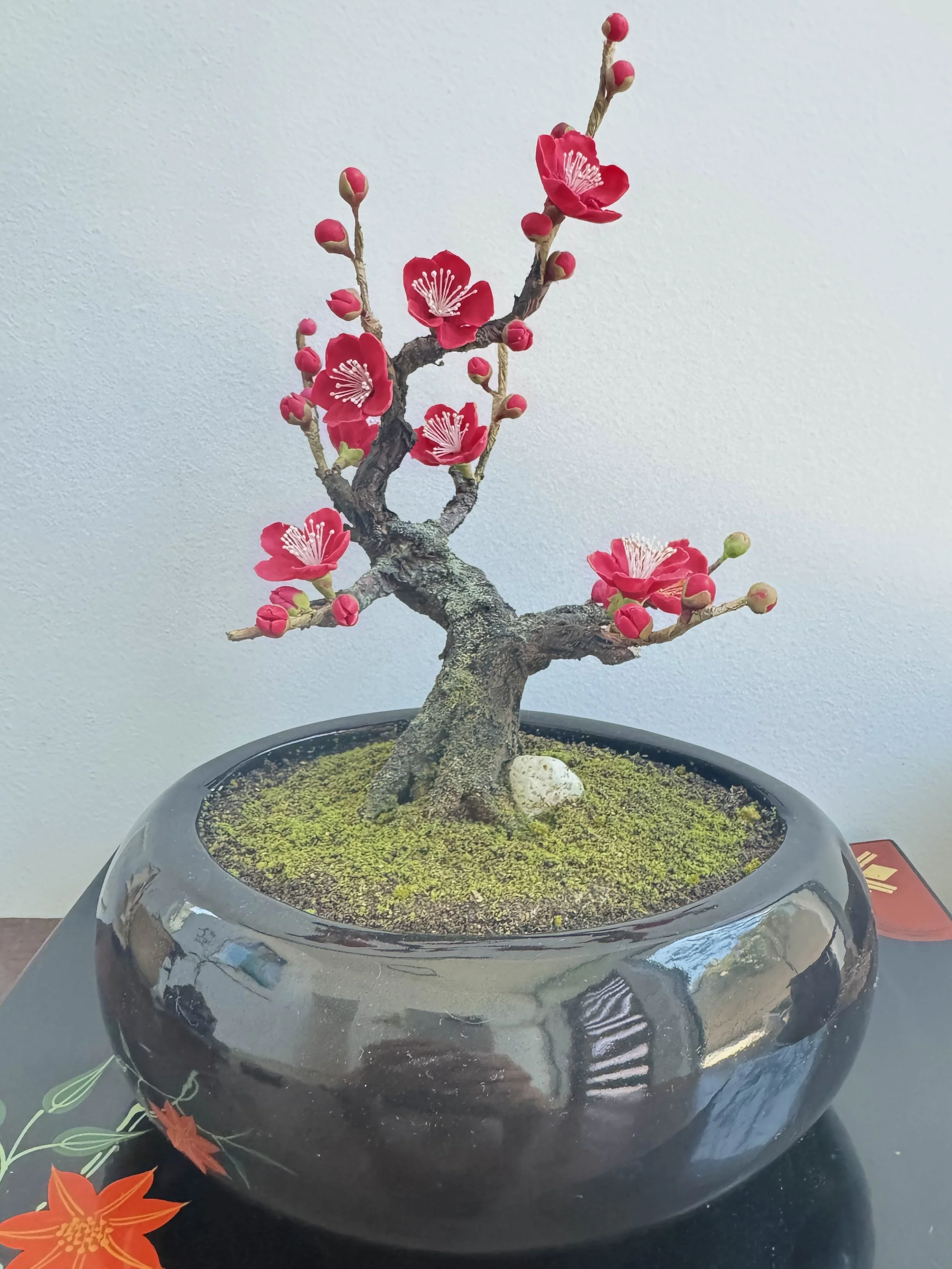 A decorative bonsai-like plant with pink flowers in a black pot, placed on a table against a plain wall.