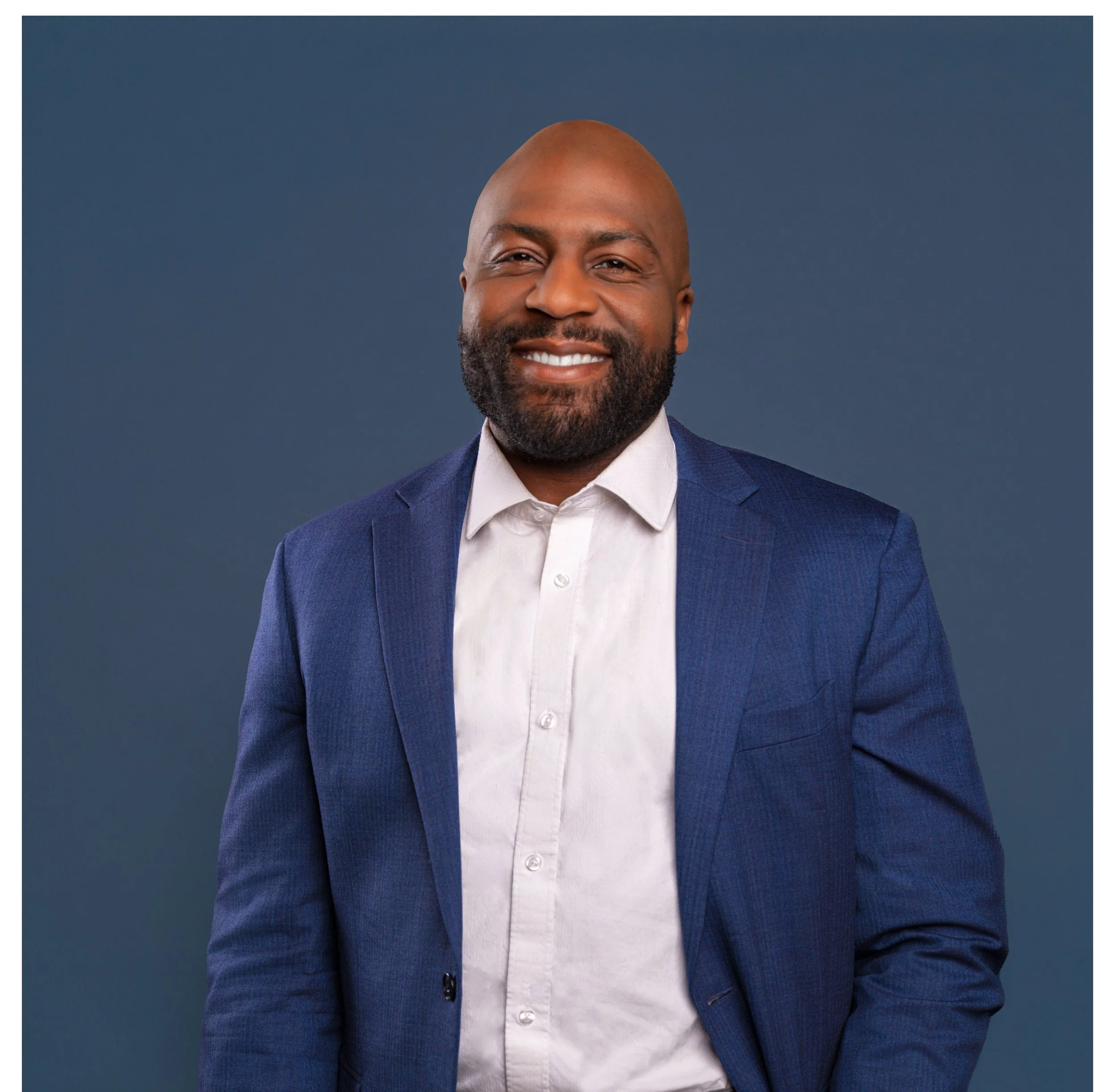 A man with a warm smile and a beard, wearing a white shirt and a blue blazer, standing against a solid dark background. Looks like a leader.