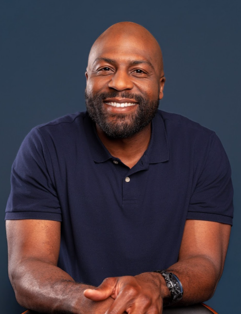 A man with a warm smile, a beard and a shaved head, wearing a navy polo shirt, sitting against a dark blue background.